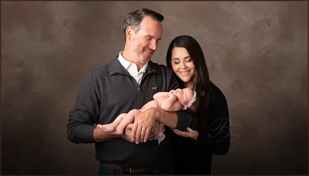 Mother and father holding newborn during Houston portrait session Mother and father holding their newborn together in a warm-toned studio portrait