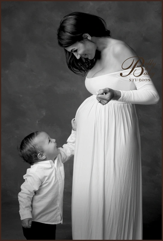 xpecting mother in a white dress looking down at her young son in a black and white maternity portrait Expecting mother in a white dress looking down at her young son in a black and white maternity portrait